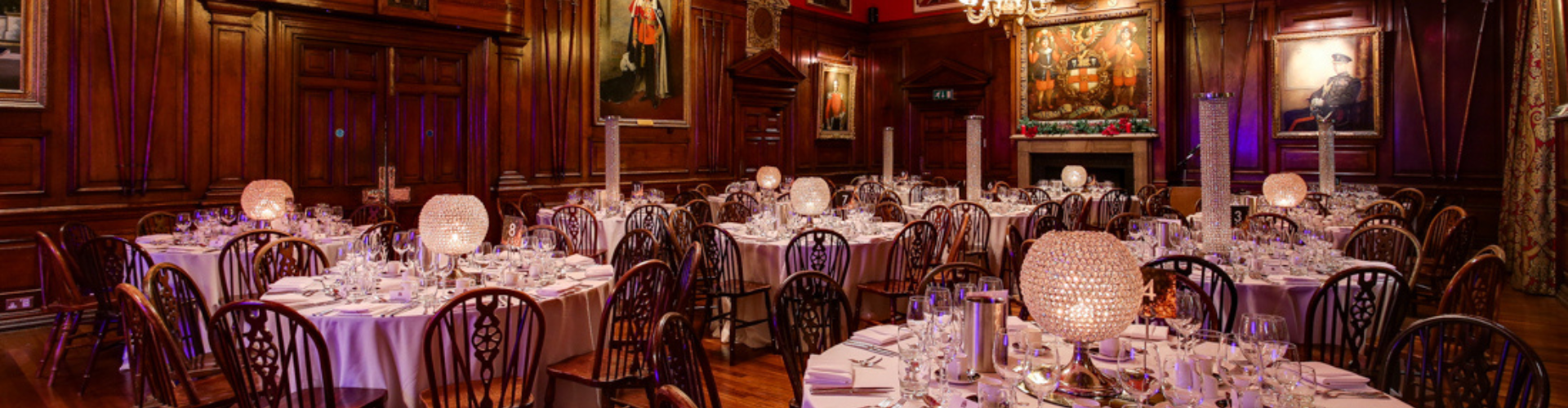 An elegant wood-panelled dining room set up for a formal event, with round tables covered in white tablecloths, arranged with glassware and illuminated centrepieces