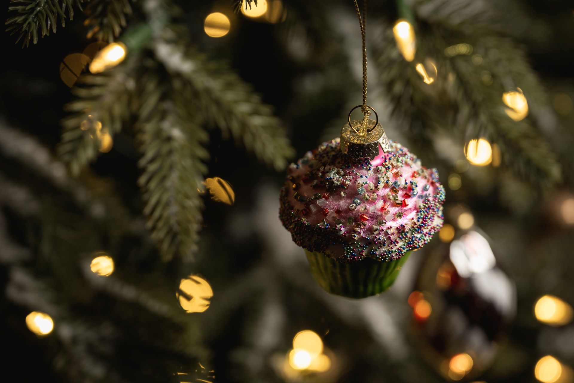 A cupcake-shaped Christmas tree ornament decorated with pink, white, and purple sprinkles, hanging from a branch with warm fairy lights in the background