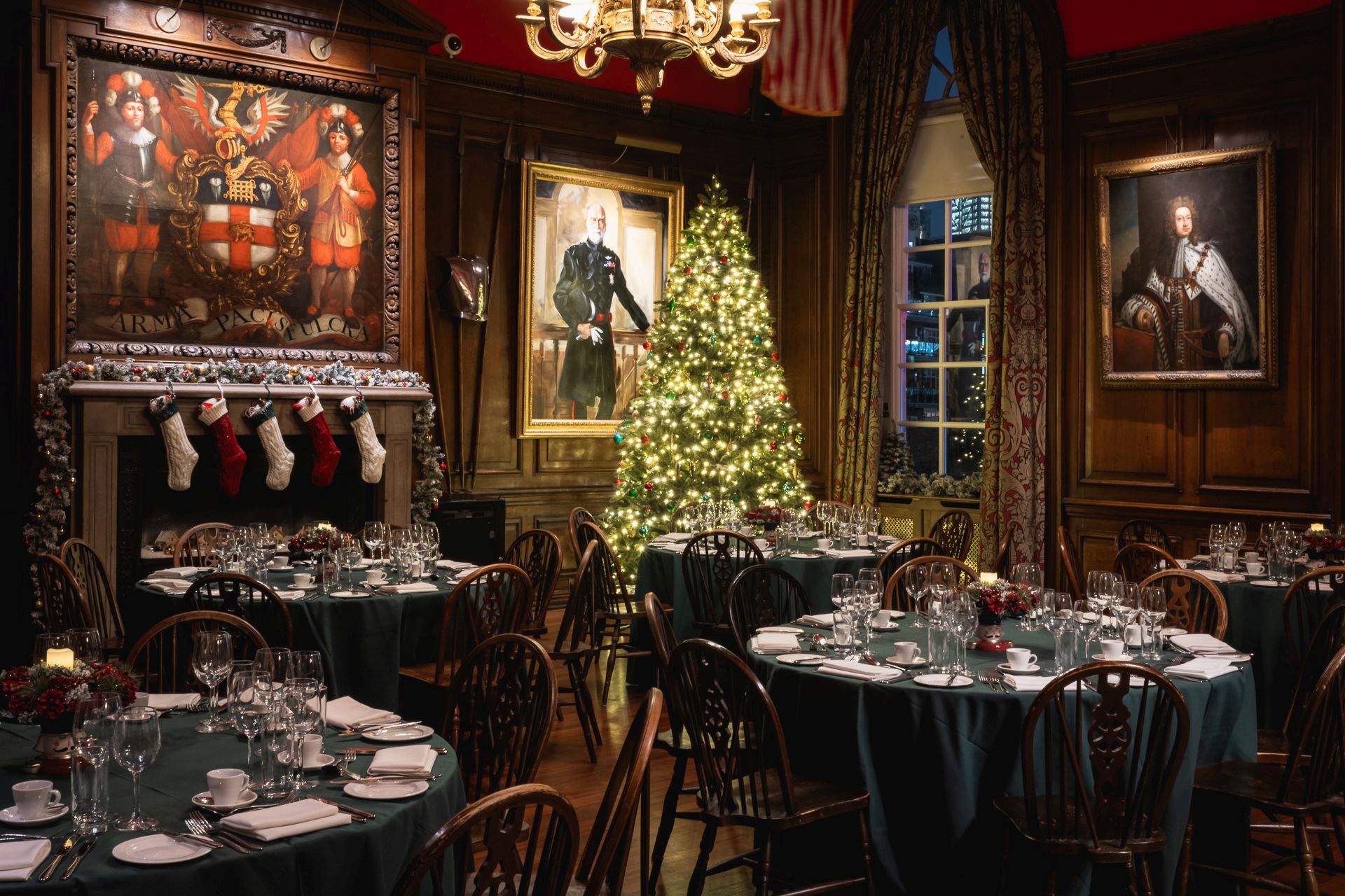 A festive dining room with round tables set for a meal, featuring a decorated Christmas tree, stockings on the fireplace, and historic portraits on wood-panelled walls