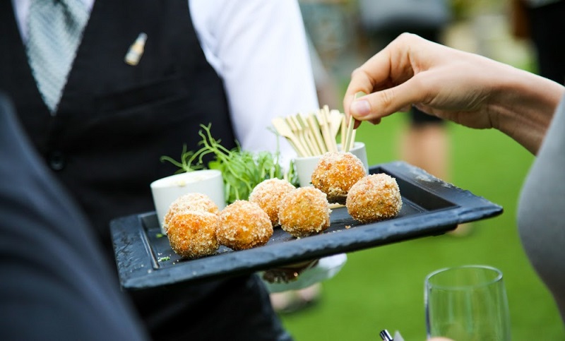 A close-up of a tray of gourmet canapés being served at an outdoor event, with a hand reaching to take one and a server in formal attire in the background.